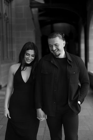 A couple in coordinated black outfits shares a tender moment while walking along a historic building.