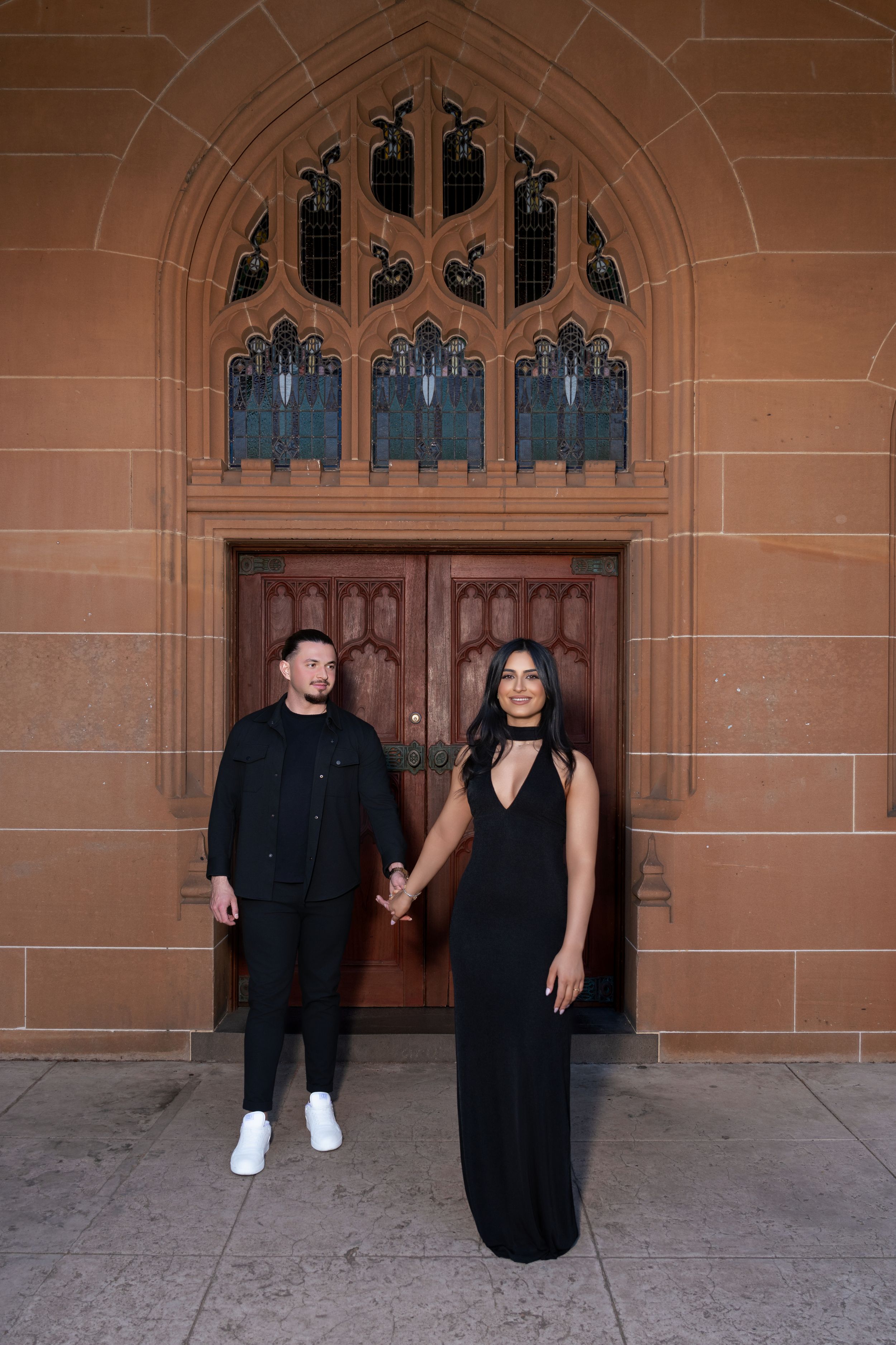 A couple poses in front of an ornate gothic doorway wearing matching black outfits.