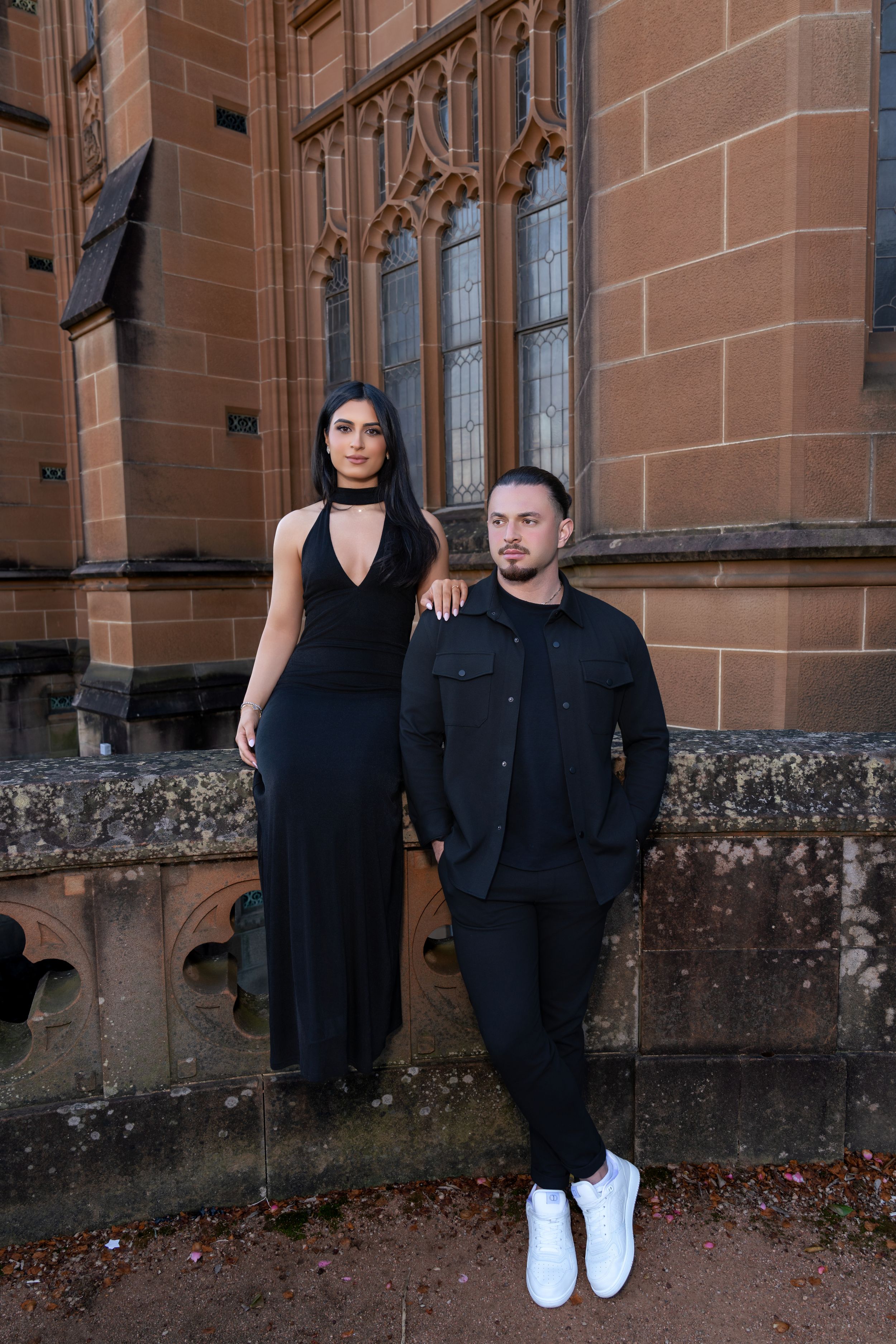 A couple in black attire poses against a sandstone church wall with gothic windows.
