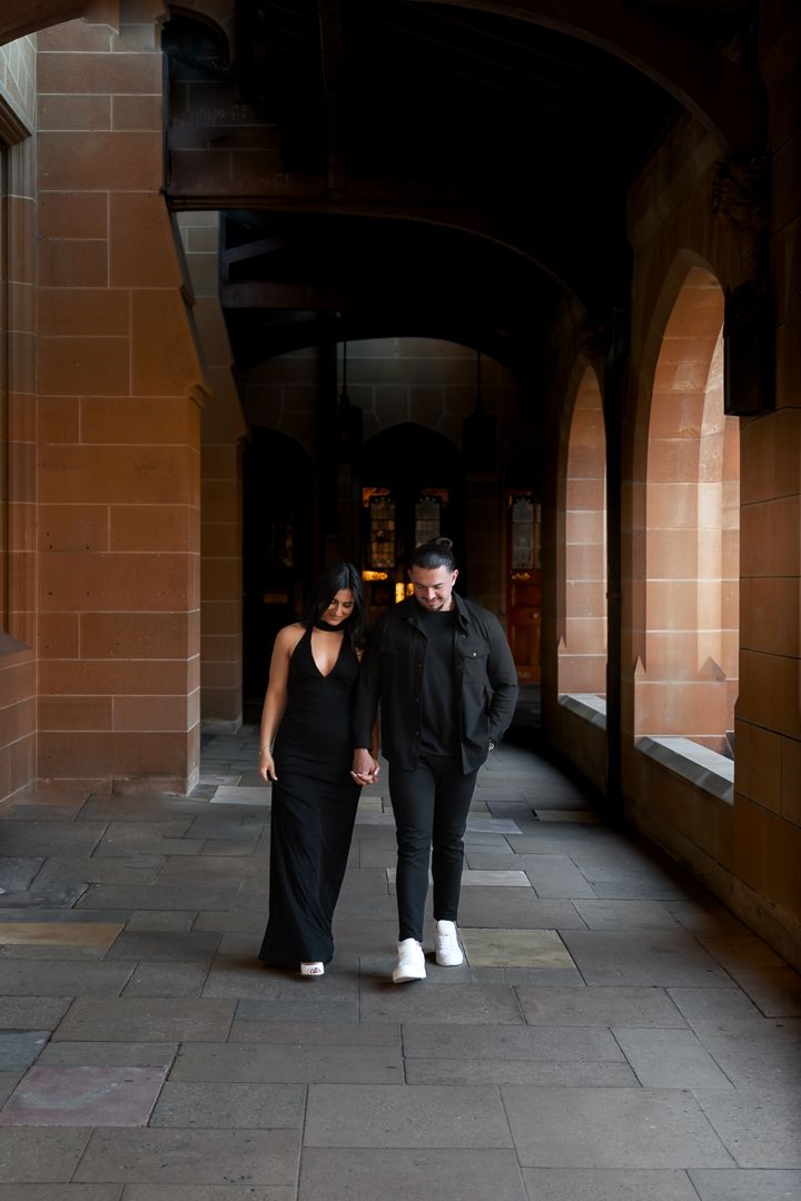 A couple walks through a dimly lit stone archway corridor in matching dark outfits.