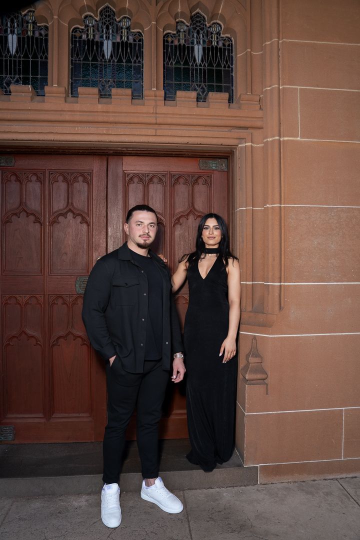A couple dressed in black stands against ornate wooden church doors and sandstone walls.