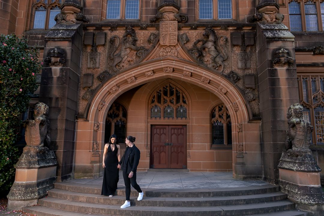 A couple in black clothing stands on stone steps leading to a gothic church entrance.