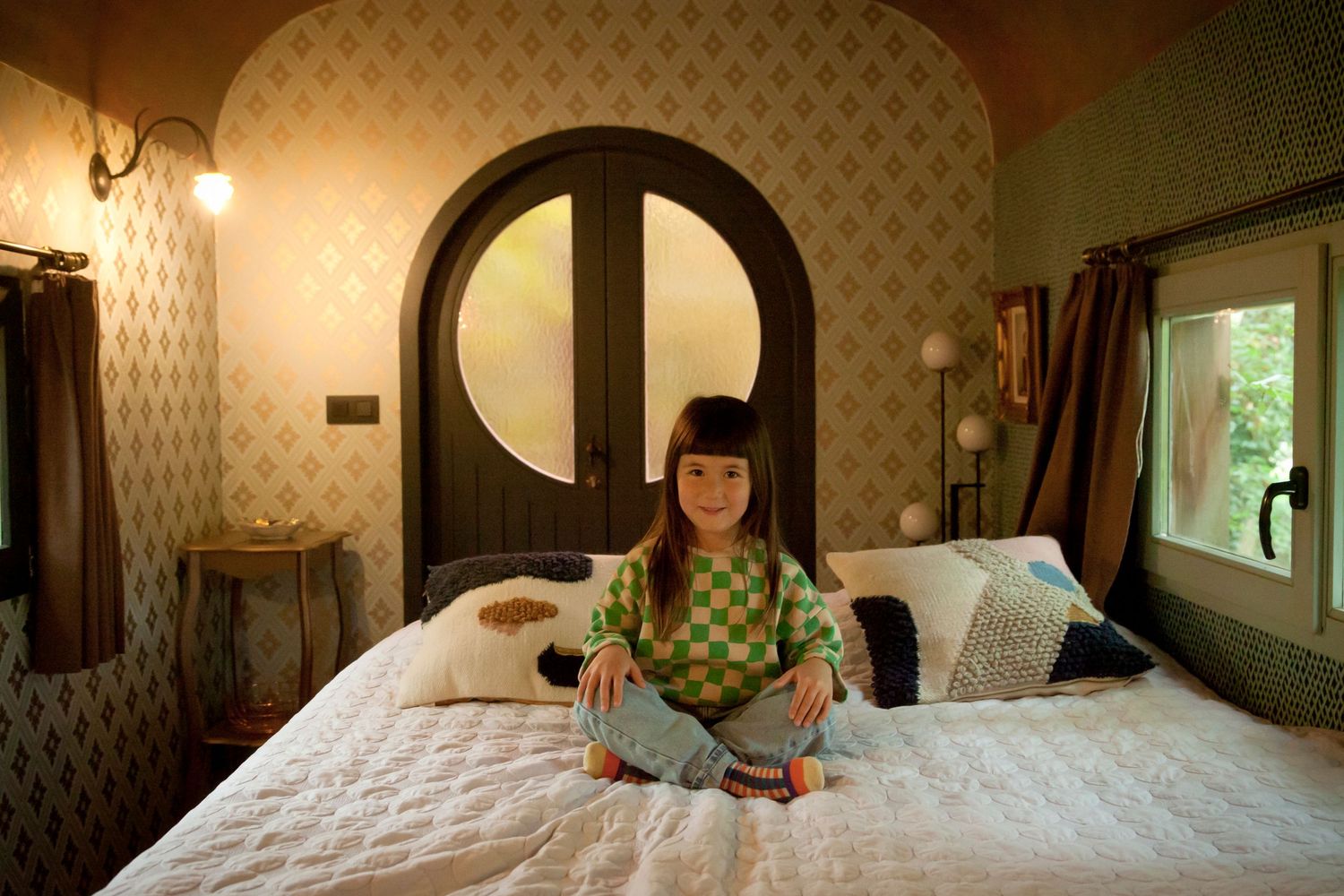 A cozy bedroom with patterned wallpaper features a white quilted bed and round arched doorway in vintage style decor.