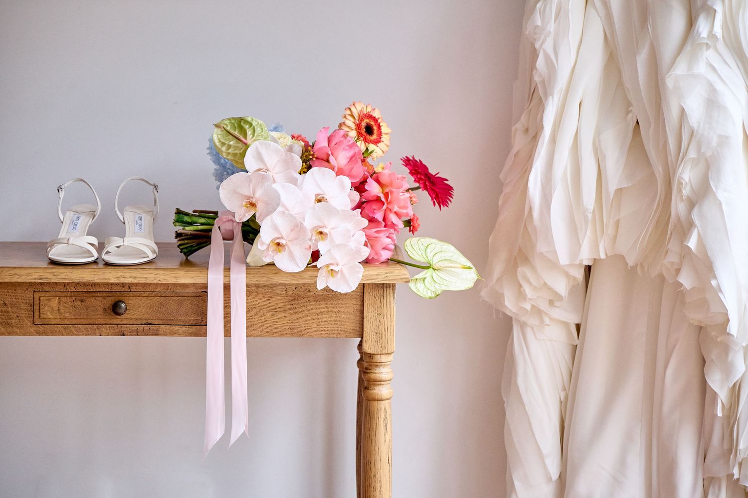 Wedding details with pink and white floral bouquet, satin ribbons, and bridal shoes on wooden table next to white dress.