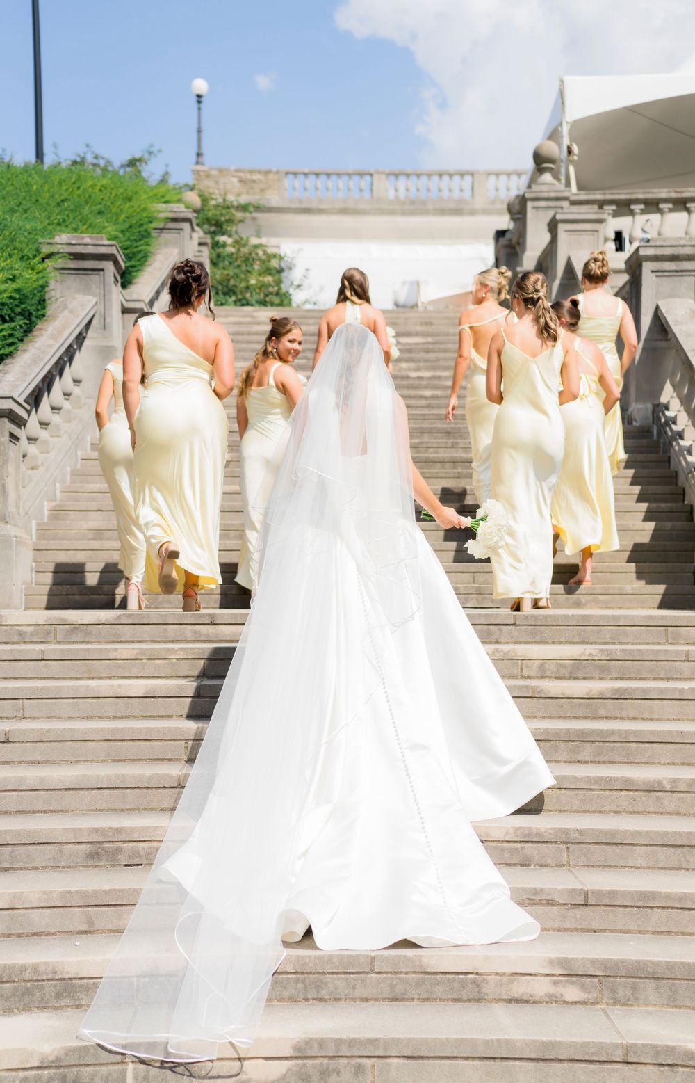 Wedding party in cream and white dresses descending elegant stone steps outdoors.