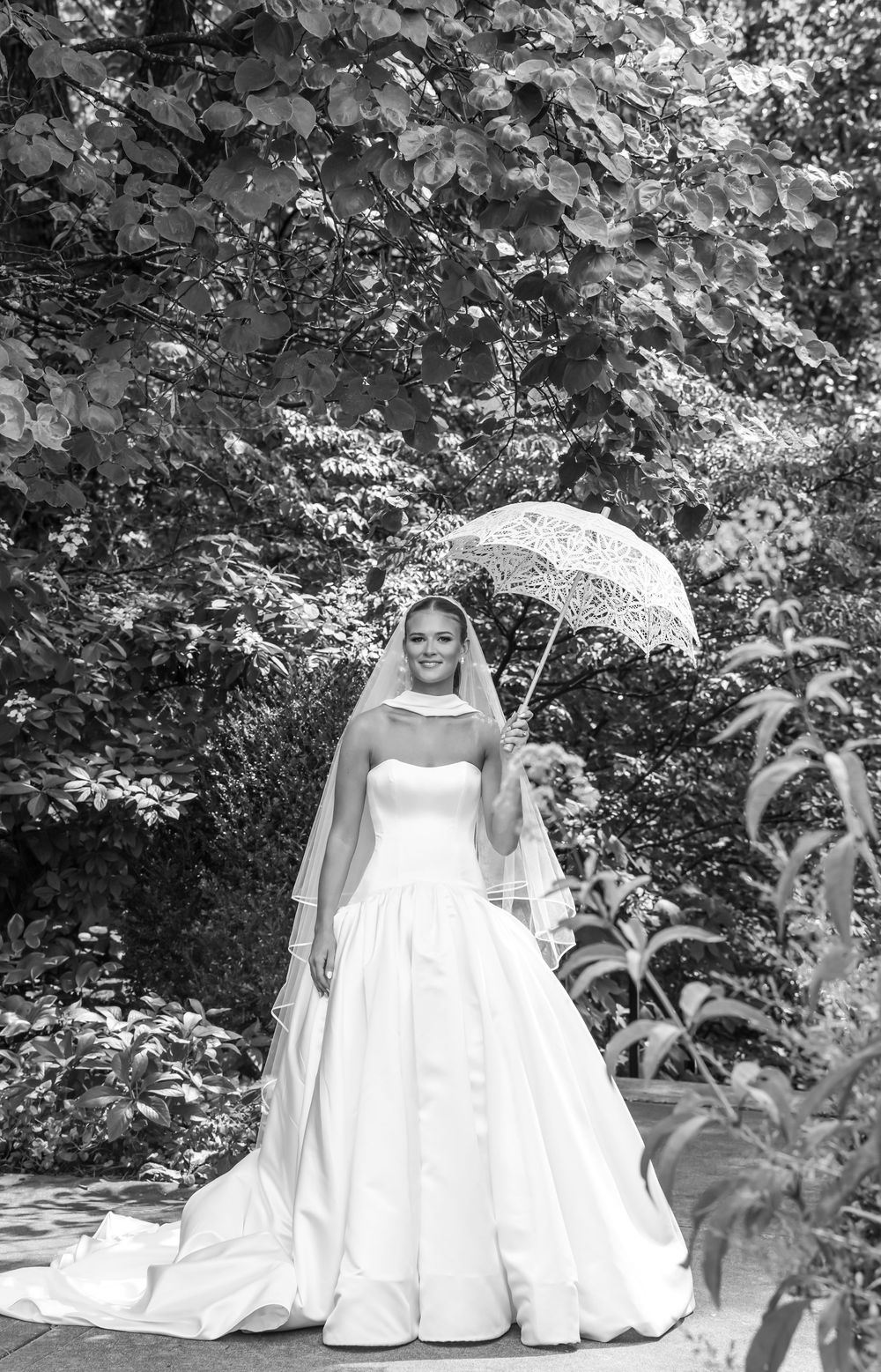 Three figures in elegant white gowns sit gracefully with parasols in a lush garden setting.