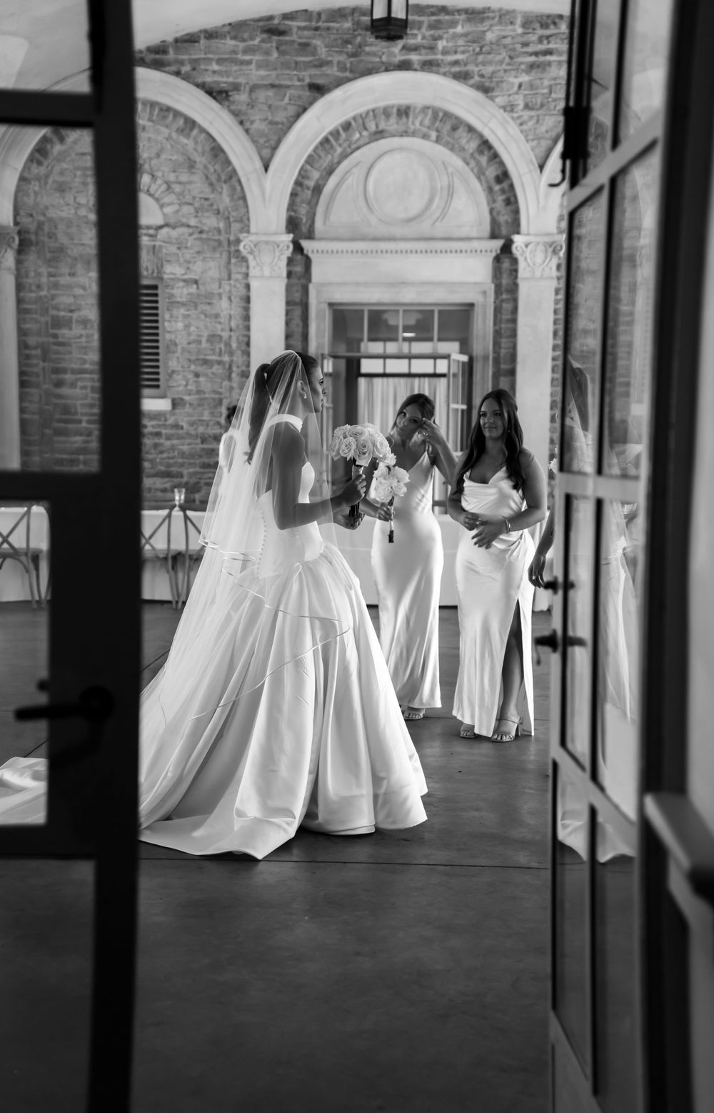 Black and white image of multiple wedding dresses displayed in an elegant brick archway corridor.