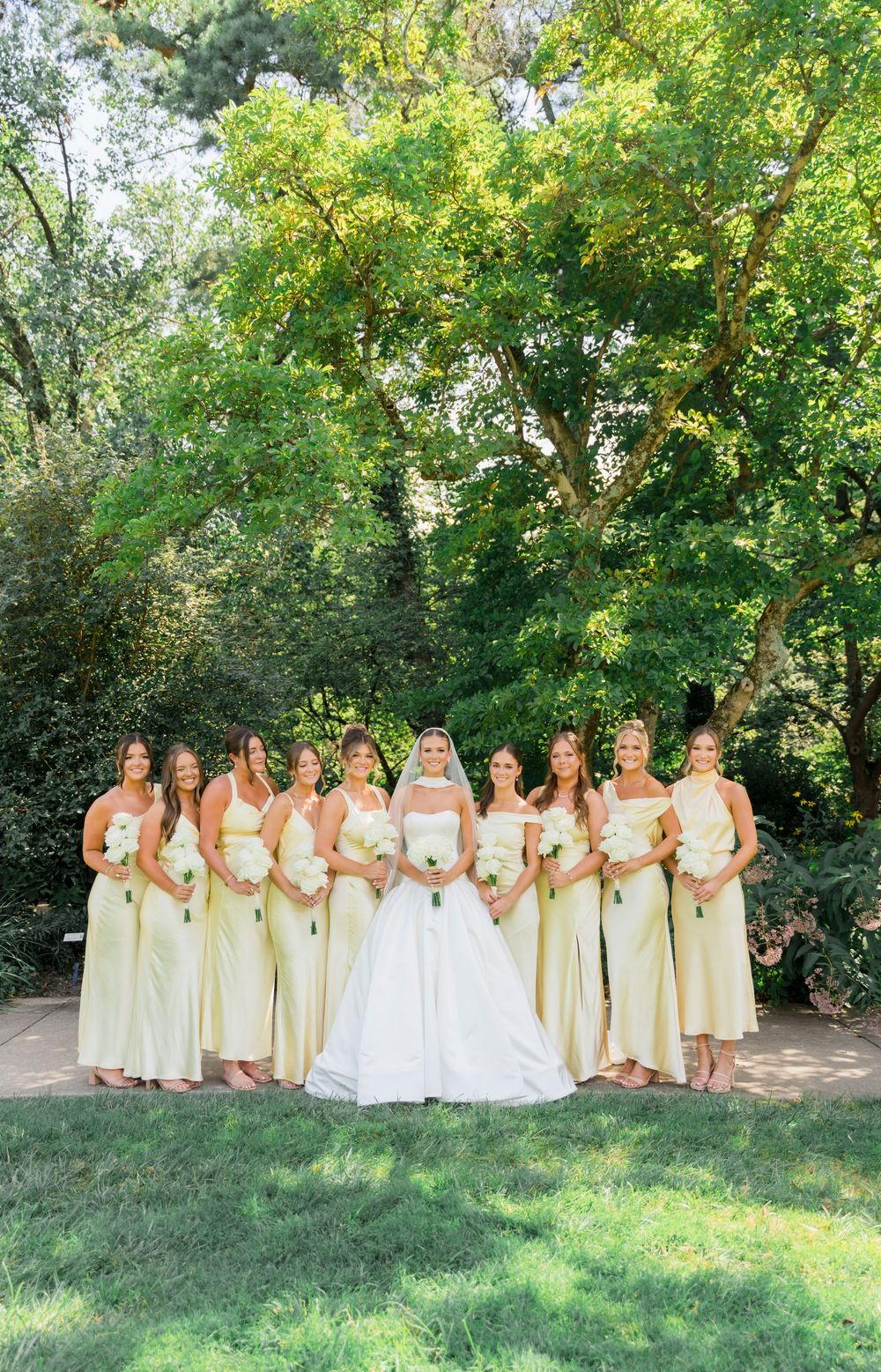 A wedding party in yellow dresses poses on a grassy hill under green trees on a sunny day.