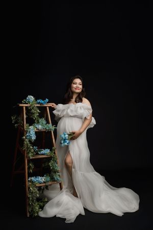 Ethereal maternity portraits in flowing white gown with blue floral ladder prop against black backdrop.