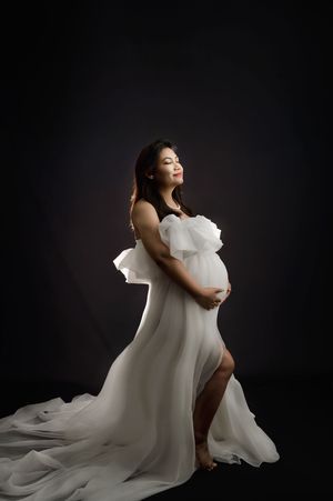 A figure in a flowing white dress poses gracefully against a dark background in an artistic maternity photoshoot series.