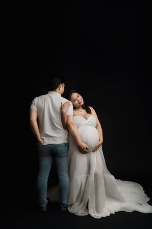 A couple in white clothing poses together against a dark backdrop for a maternity photoshoot.