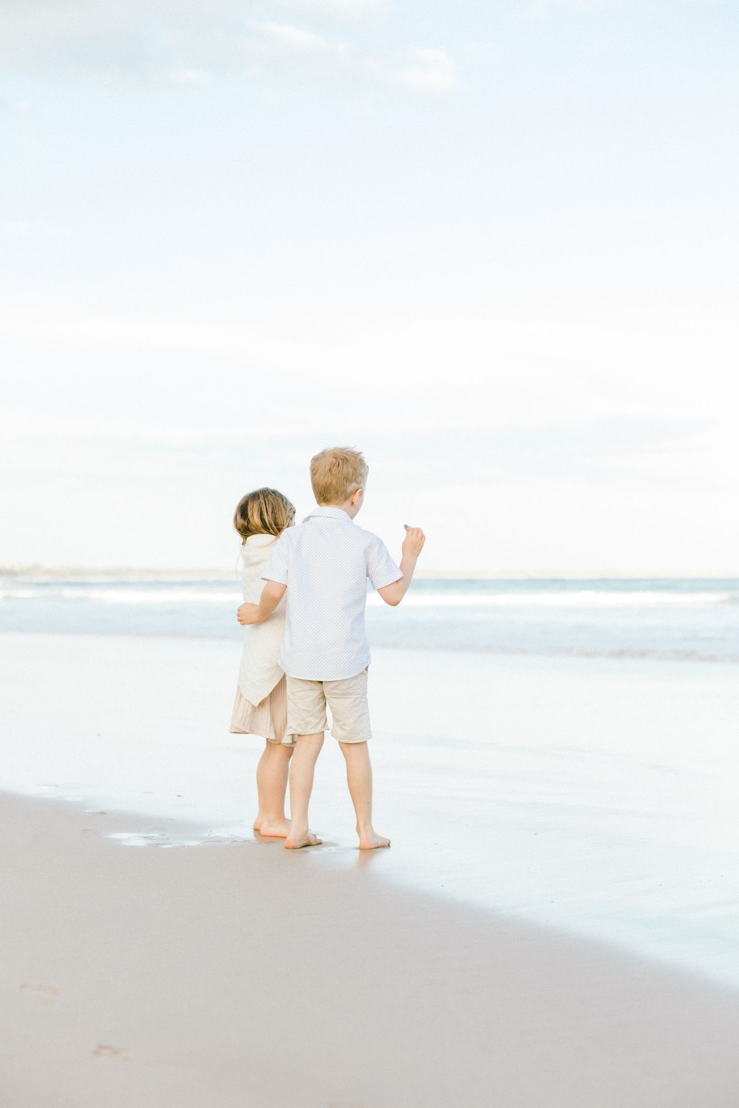 Two small children in white and beige clothing walk together along a serene beach shoreline at sunset.