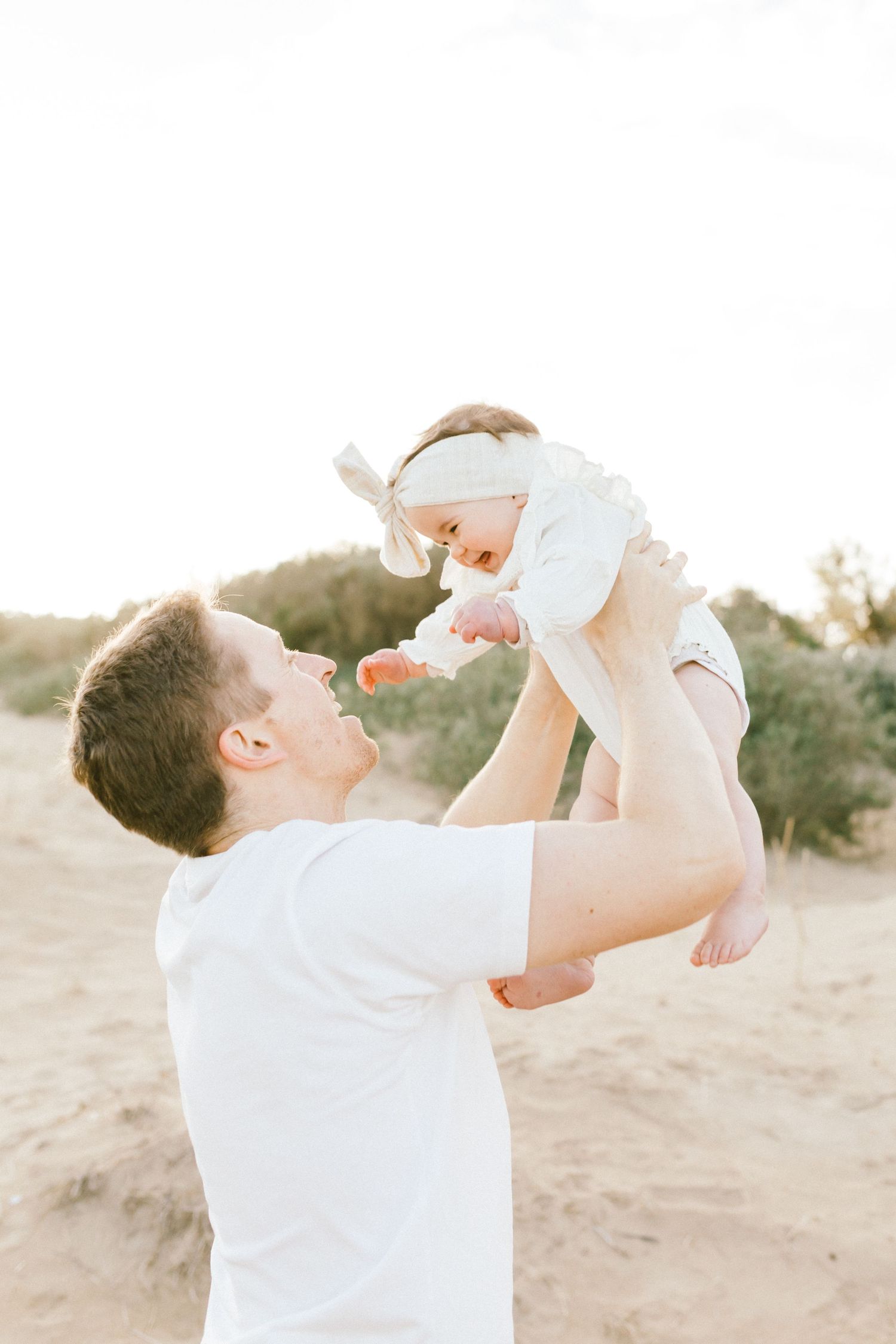 A person in white clothing lifts and plays with a baby outdoors against a natural desert background.