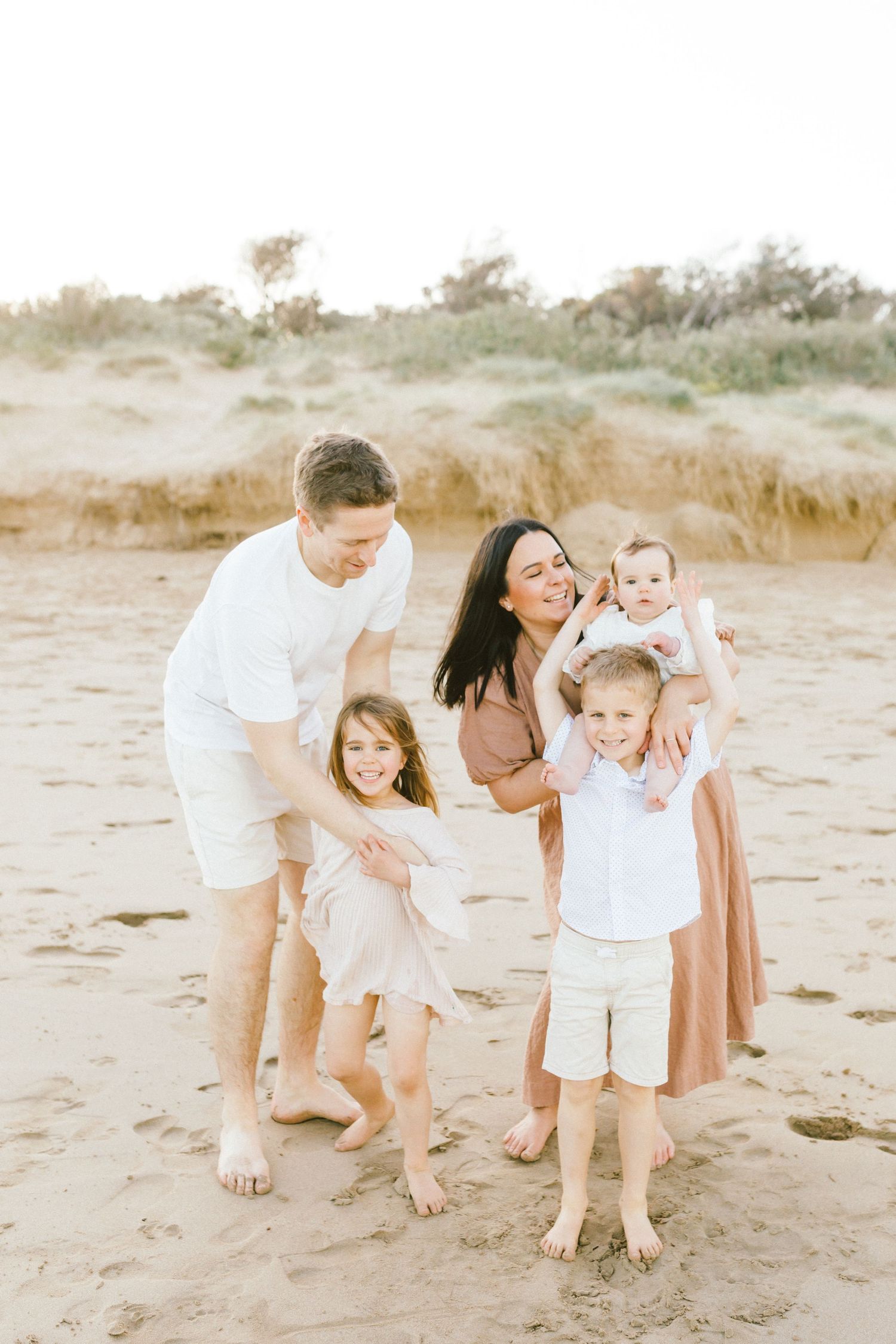 Family enjoying playful moments together on a sandy beach in soft neutral tones during sunset.