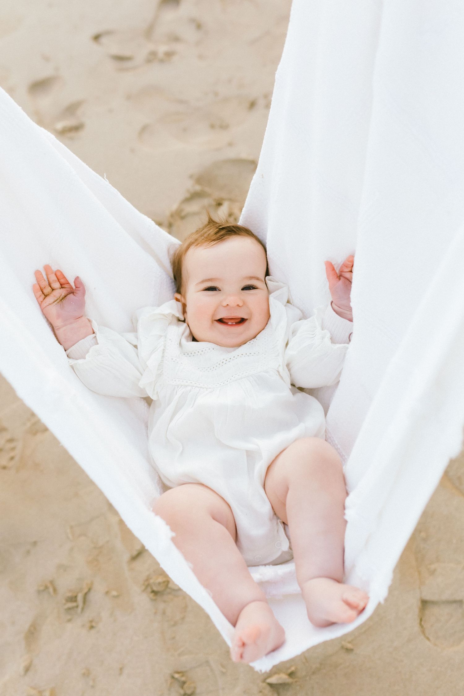 Happy baby wearing white clothing lying on flowing white fabric against a neutral background.