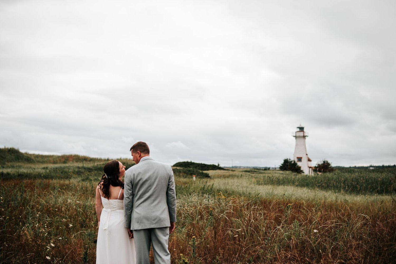 A couple stands together in a scenic meadow with wildflowers and cloudy skies during their wedding day.