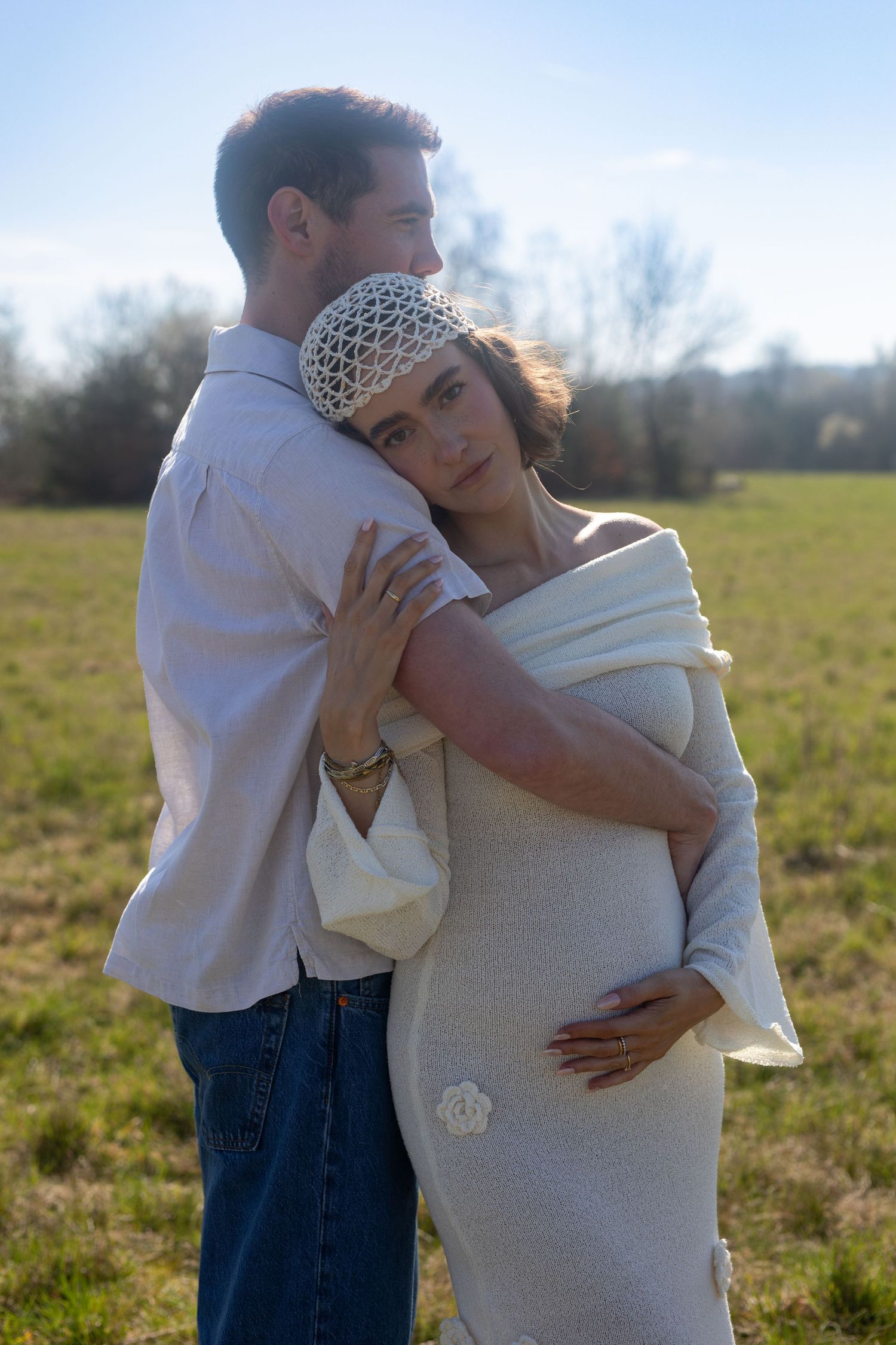 Couple embracing outdoors with pregnant woman in cream knit dress and white crochet headband.