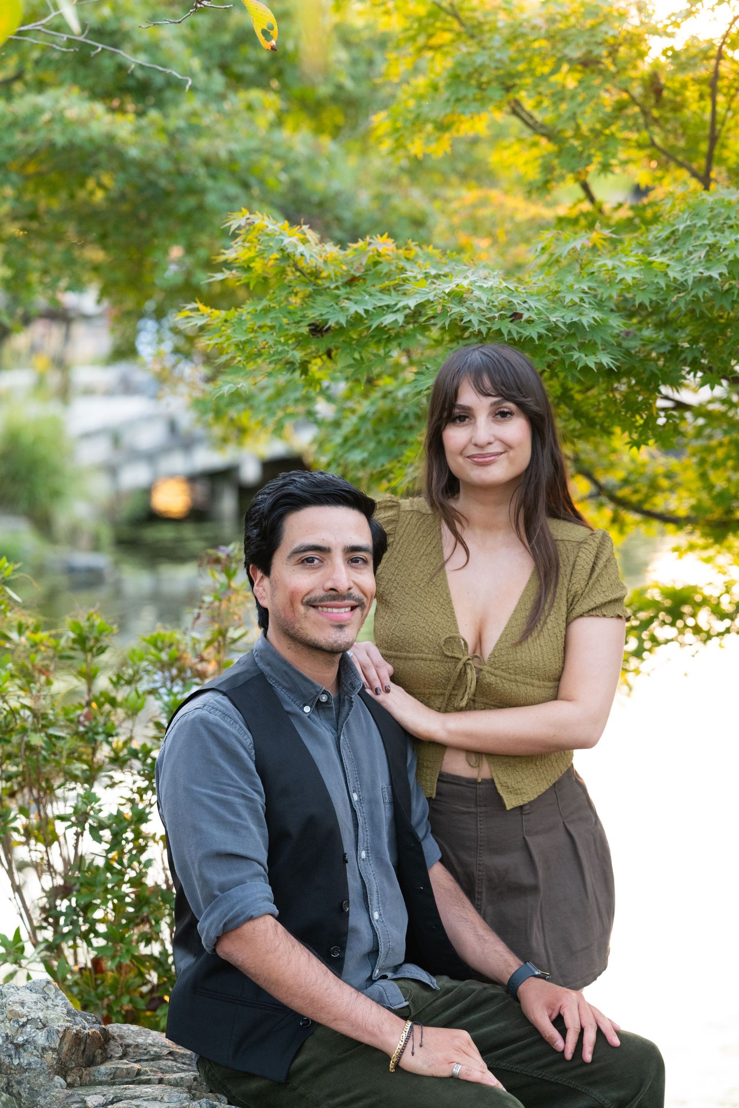 Smiling couple posing together by a tranquil pond surrounded by lush green trees.