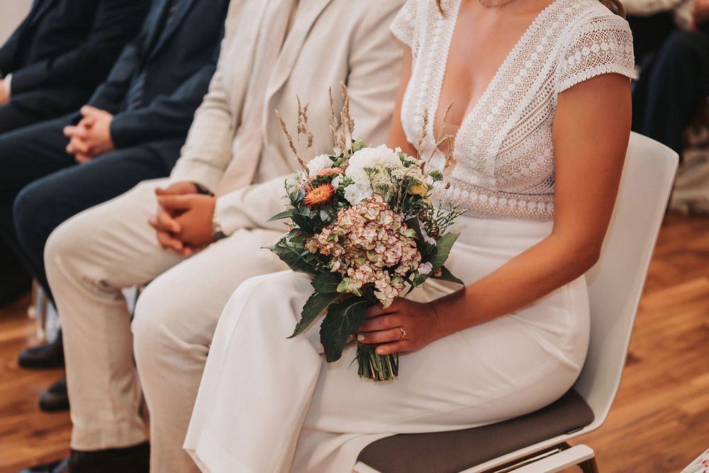 A person wearing a white wedding dress holds a pink and white floral bouquet while sitting in a chair.