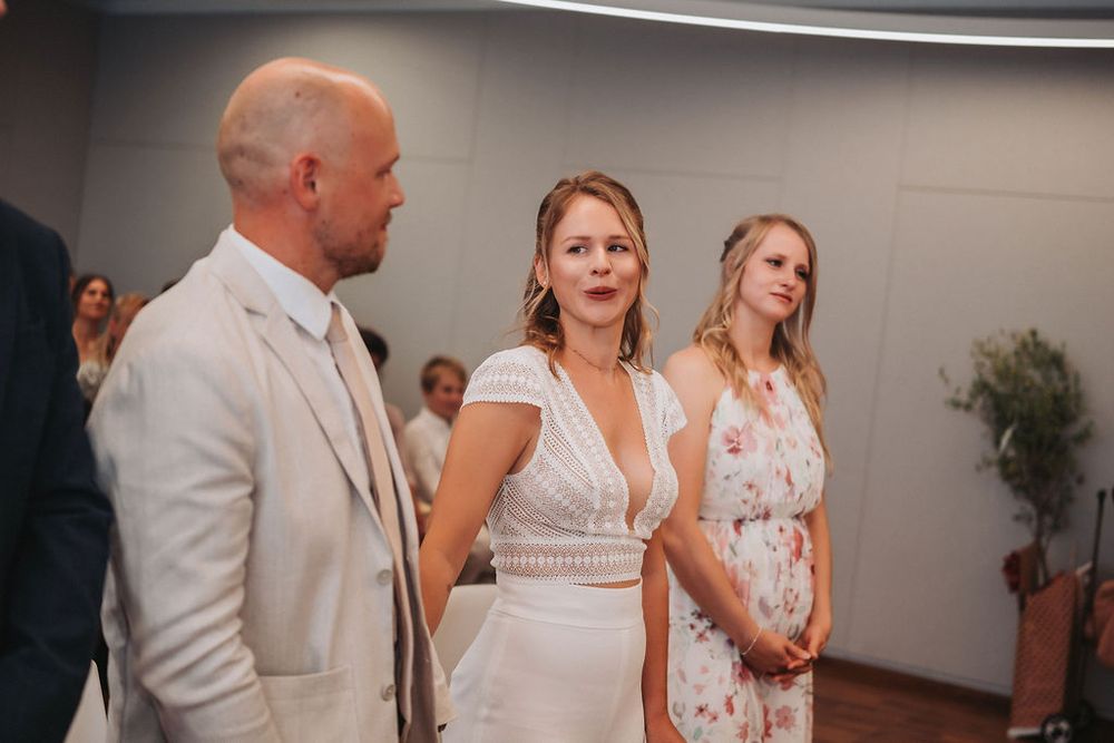 Wedding guests mingle and dance at an indoor reception venue with white walls and ambient lighting.