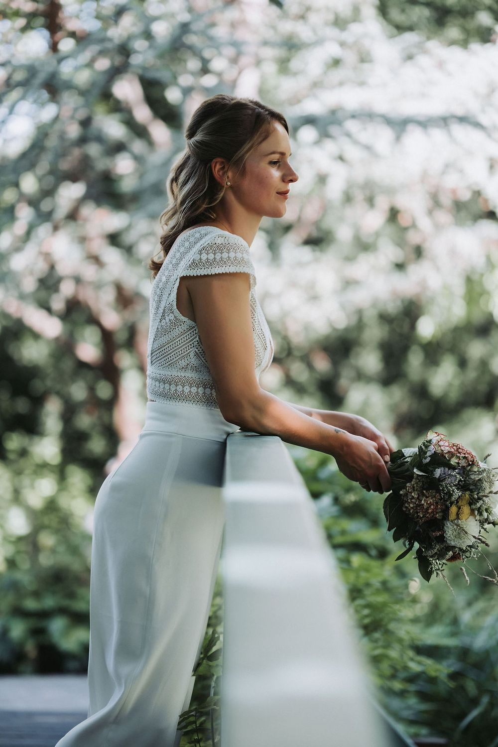 Side profile of bride in elegant white lace dress holding rustic bouquet in outdoor garden setting.