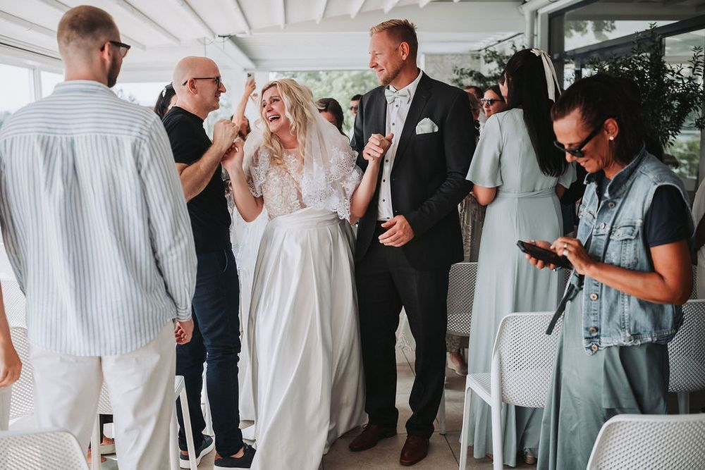 Wedding guests in white and dark attire gather and socialize during a celebration at an indoor venue.