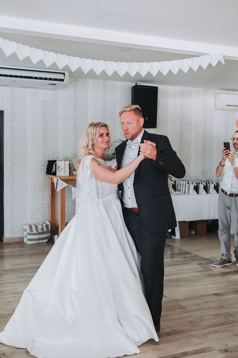 A couple dances together in a bright indoor venue with white decor and hardwood floors during their wedding celebration.