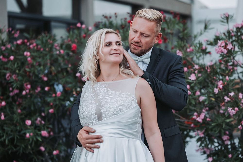 A romantic wedding couple embraces outdoors surrounded by blooming pink flowers and greenery.
