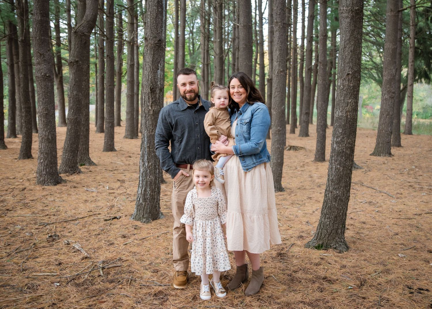 A family poses together in a serene pine forest setting wearing coordinated neutral-toned outfits.
