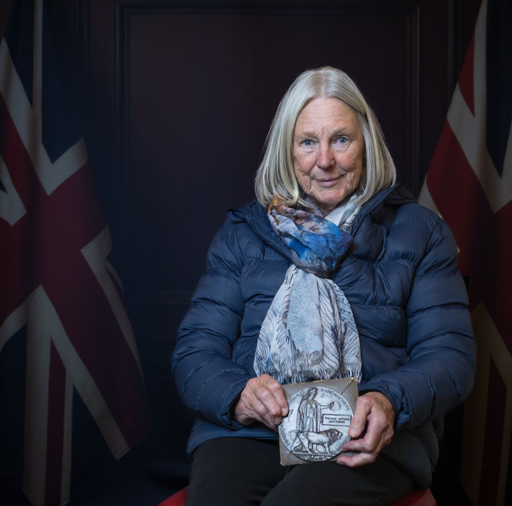 Person in a navy blue winter coat and light scarf sits against dark background with flags visible.