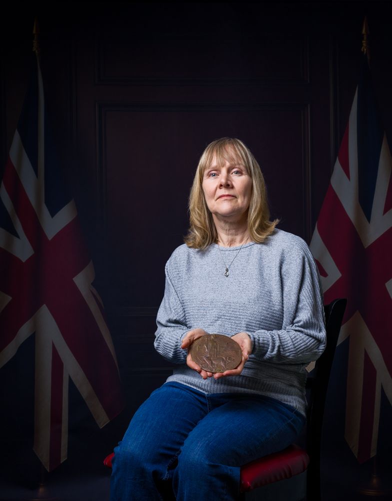 A person in a gray sweater and blue jeans sits with a Union Jack flag backdrop in a dark studio setting.