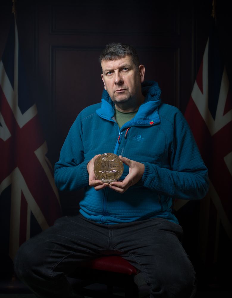 A person in a blue jacket holds a medal while seated in front of Union Jack flags in a dark setting.