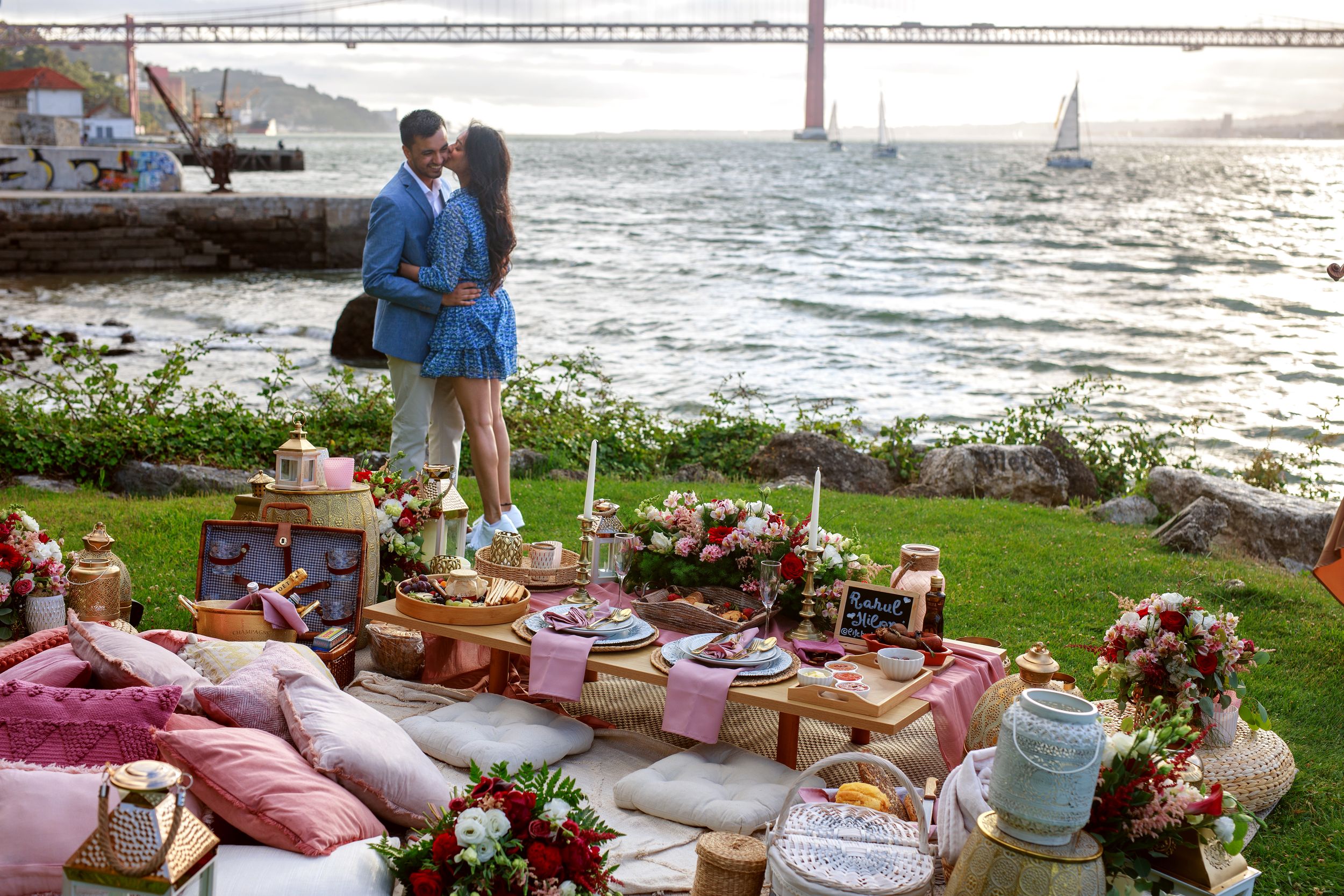 Elegant beach picnic setup with floral arrangements, pillows and decorative elements along the oceanfront.