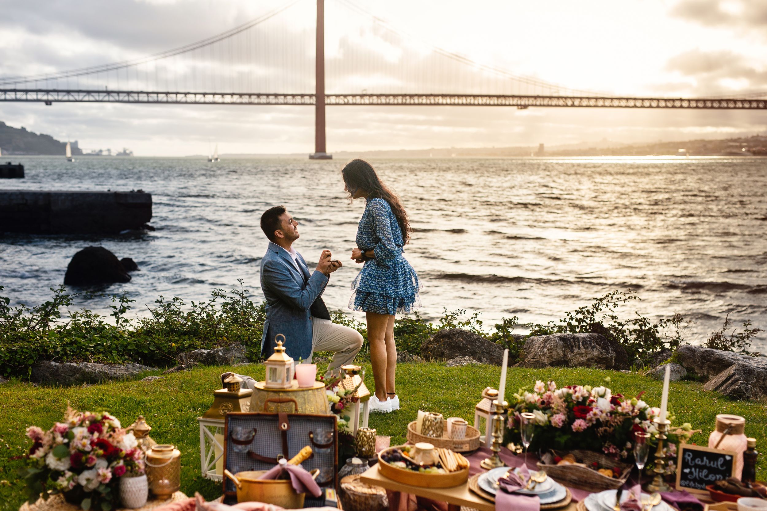 A romantic sunset picnic spread by the ocean with wine glasses, charcuterie, and flowers on a blanket.