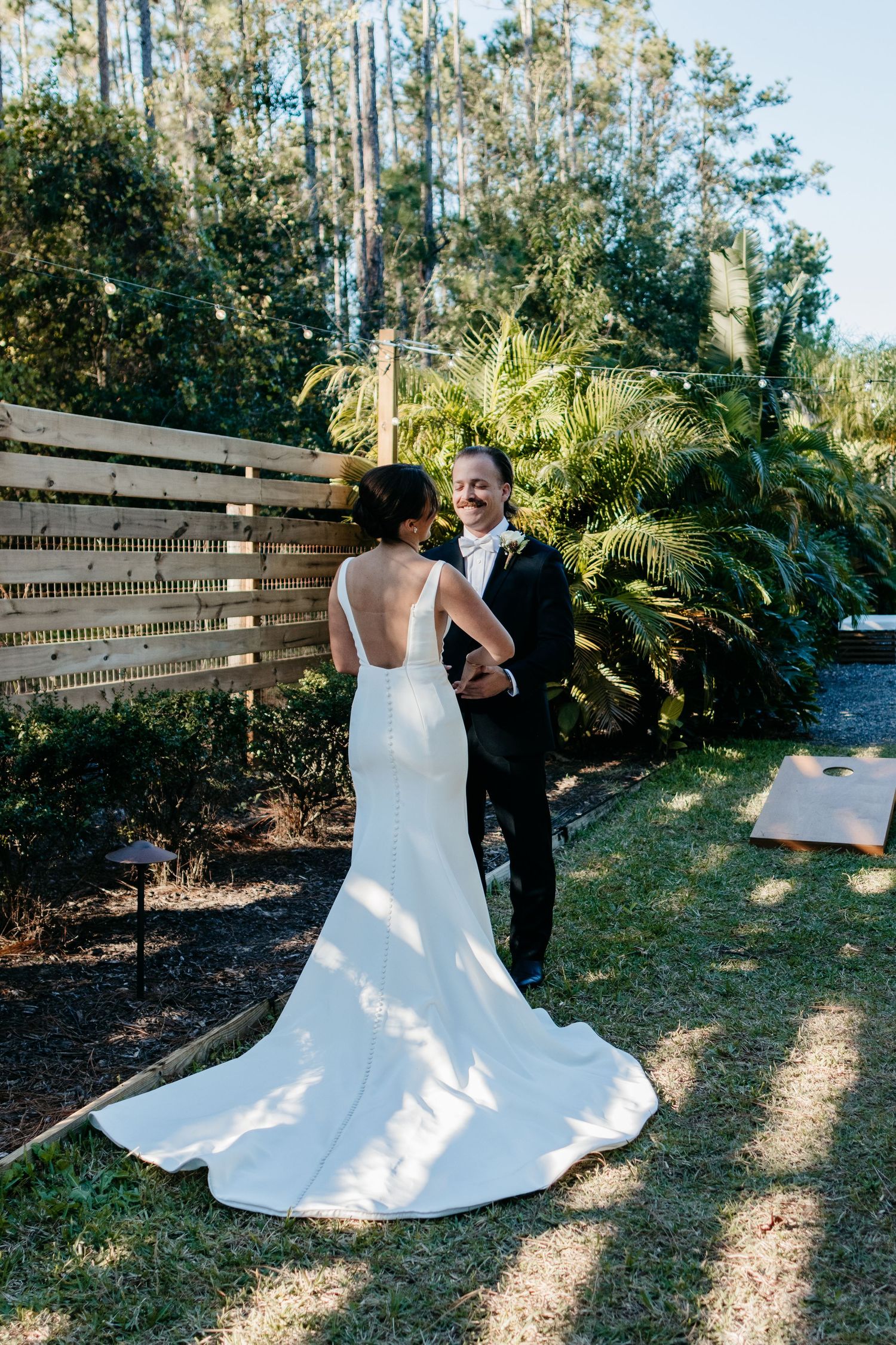 Wedding couple poses in a garden setting with tall trees and wooden fencing as a backdrop.