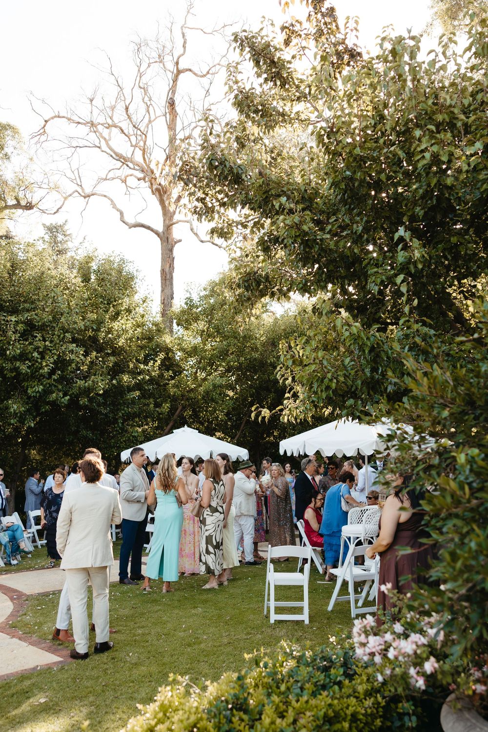 Outdoor wedding ceremony in a lush garden with guests gathered near white chairs and umbrellas beneath tall trees.
