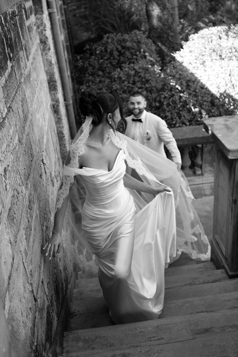 Black and white wedding photo of bride in elegant gown ascending stairs as smiling groom follows behind.