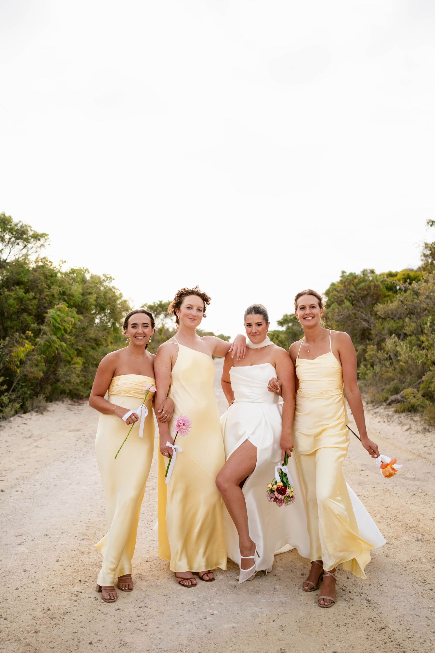 Wedding party wearing pale yellow dresses and white gown pose together on sandy beach path during outdoor celebration.