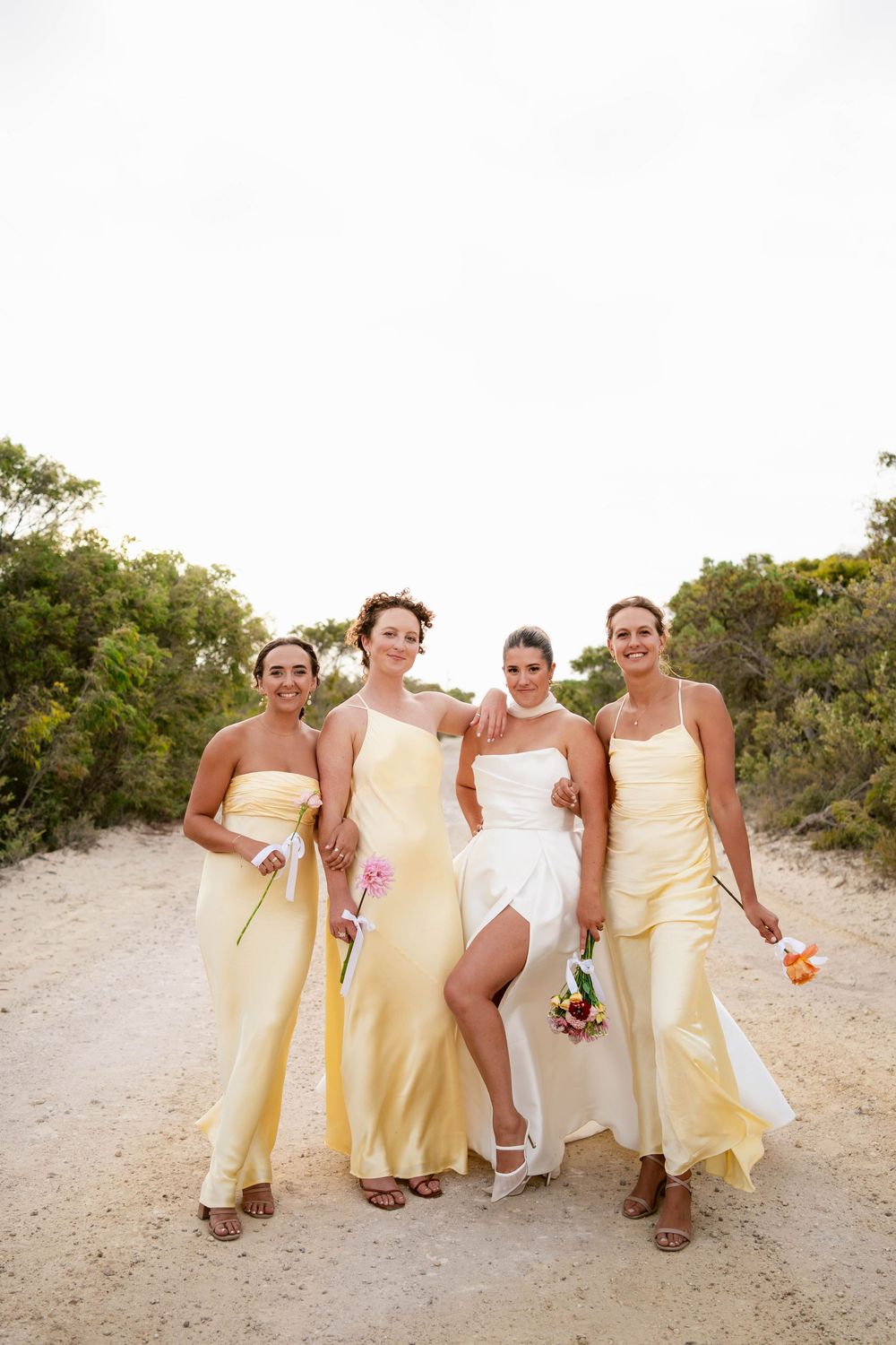 Wedding party wearing pale yellow dresses and white gown pose together on sandy beach path during outdoor celebration.