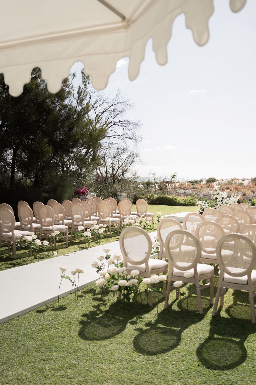 Elegant outdoor wedding ceremony setup with ivory French Louis chairs arranged in rows on green grass under a scalloped tent.