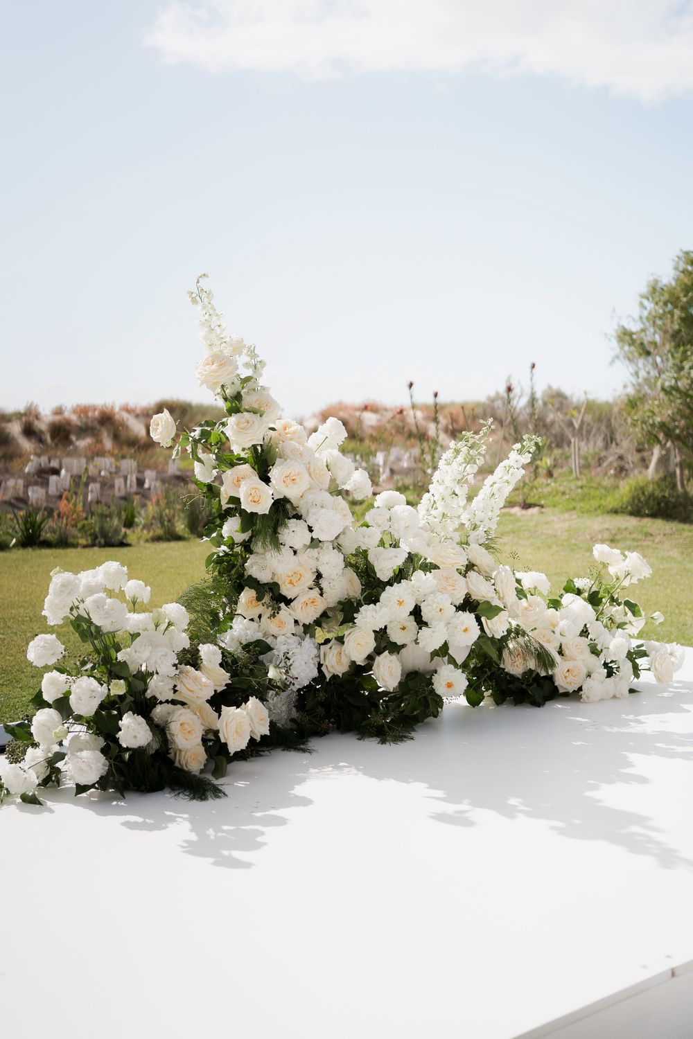 Elegant white floral arrangement with roses and greenery on a white surface at an outdoor wedding ceremony.