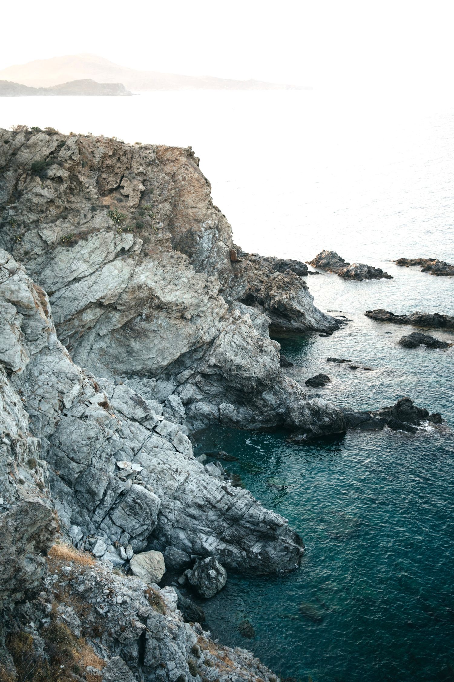 Dramatic rocky coastal cliffs tower over deep blue ocean waters against a cloudy sky.