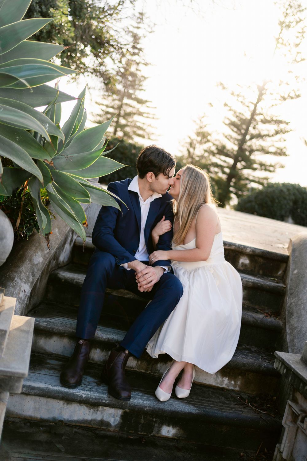 A romantic couple shares tender moments at sunset while sitting on a stone wall surrounded by lush greenery.