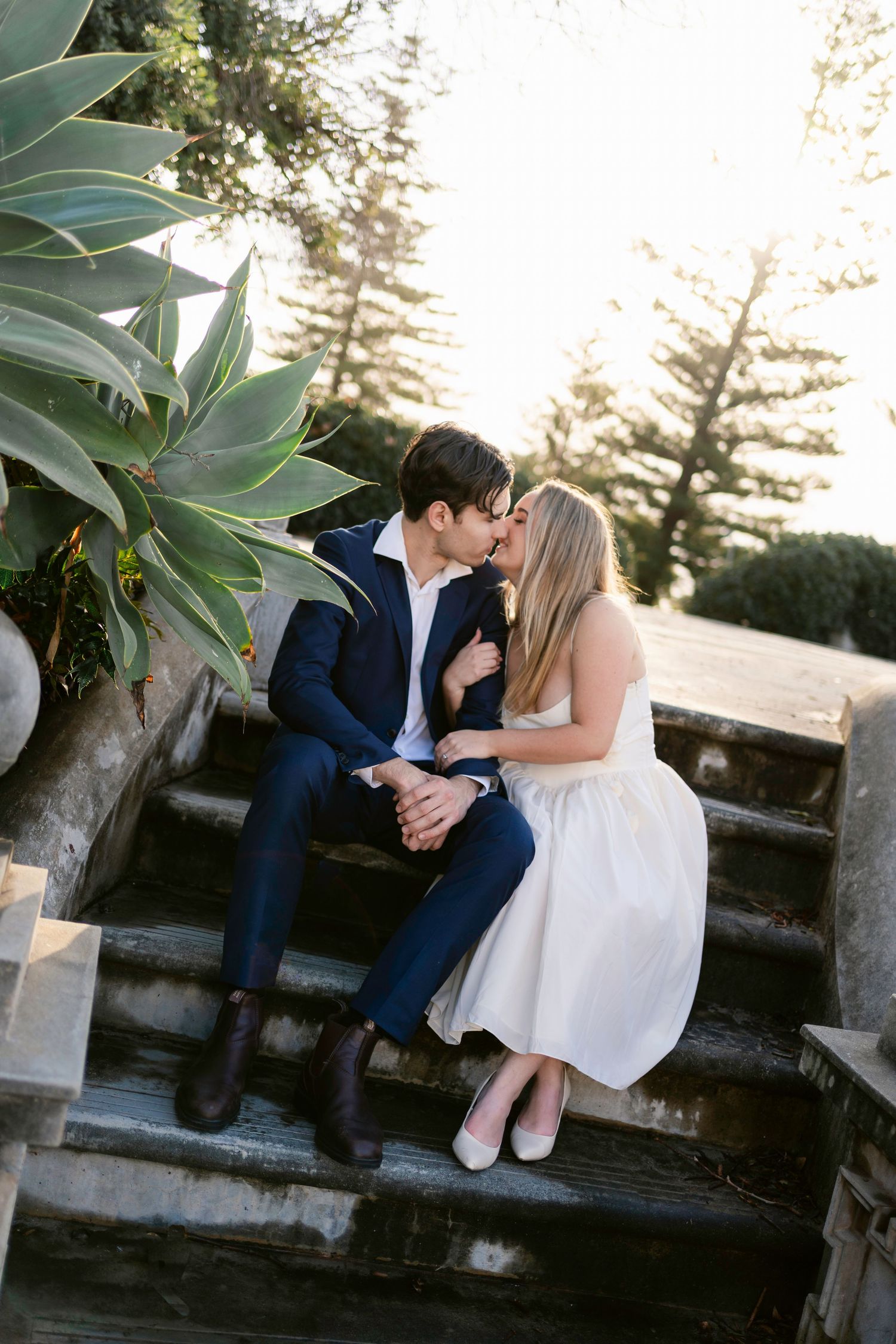 A romantic couple shares tender moments at sunset while sitting on a stone wall surrounded by lush greenery.