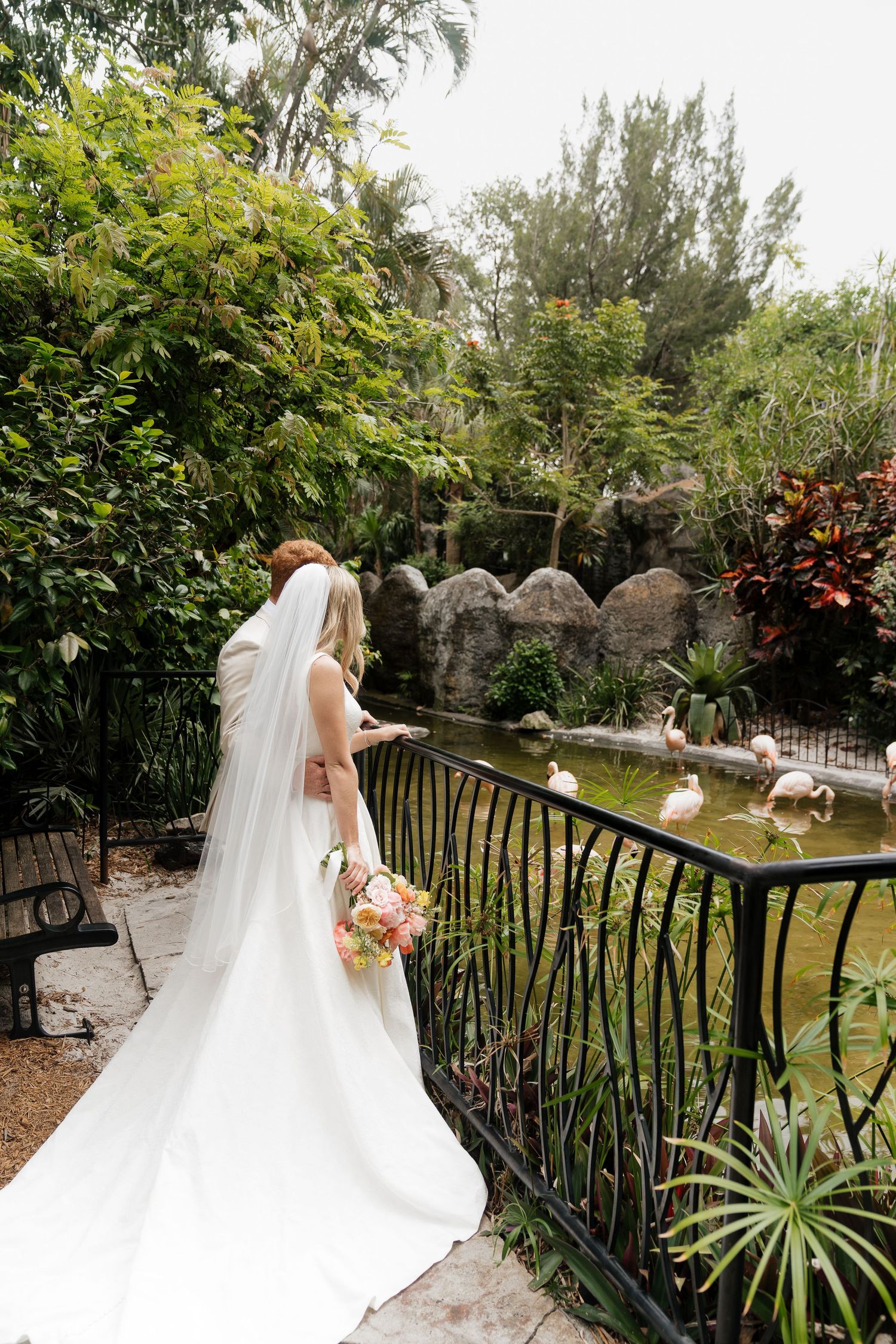 A bride in a flowing white wedding gown with a long veil stands on a garden pathway surrounded by lush greenery.
