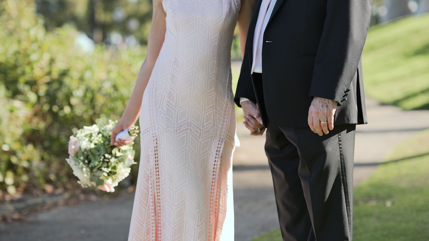 Couple in formal attire walking together on a sunny path surrounded by greenery.
