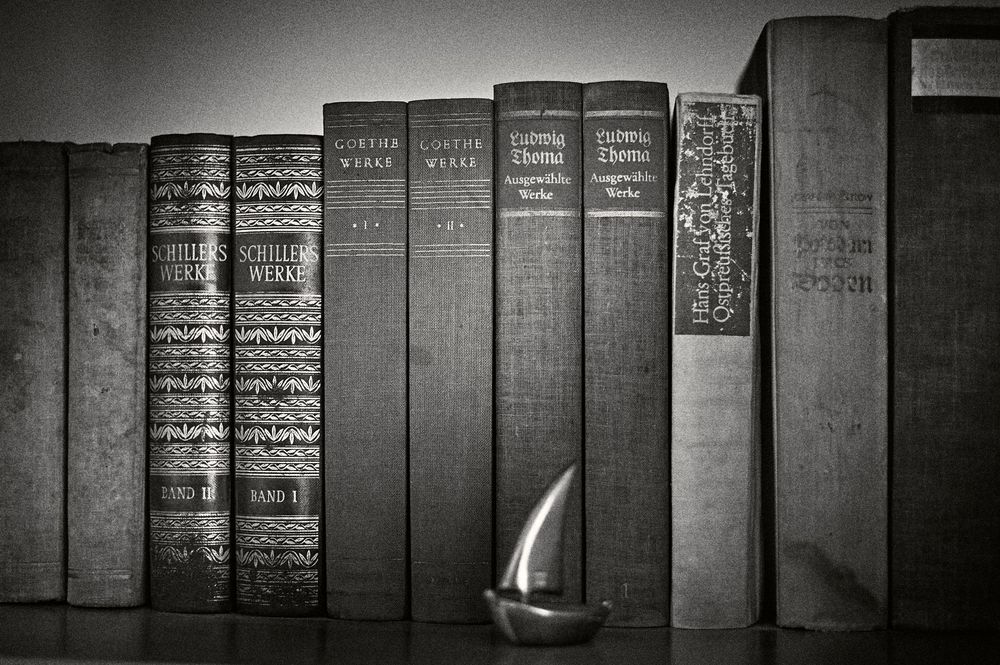 A row of vintage hardcover books stands on a shelf with a small lit candle flame glowing in black and white.
