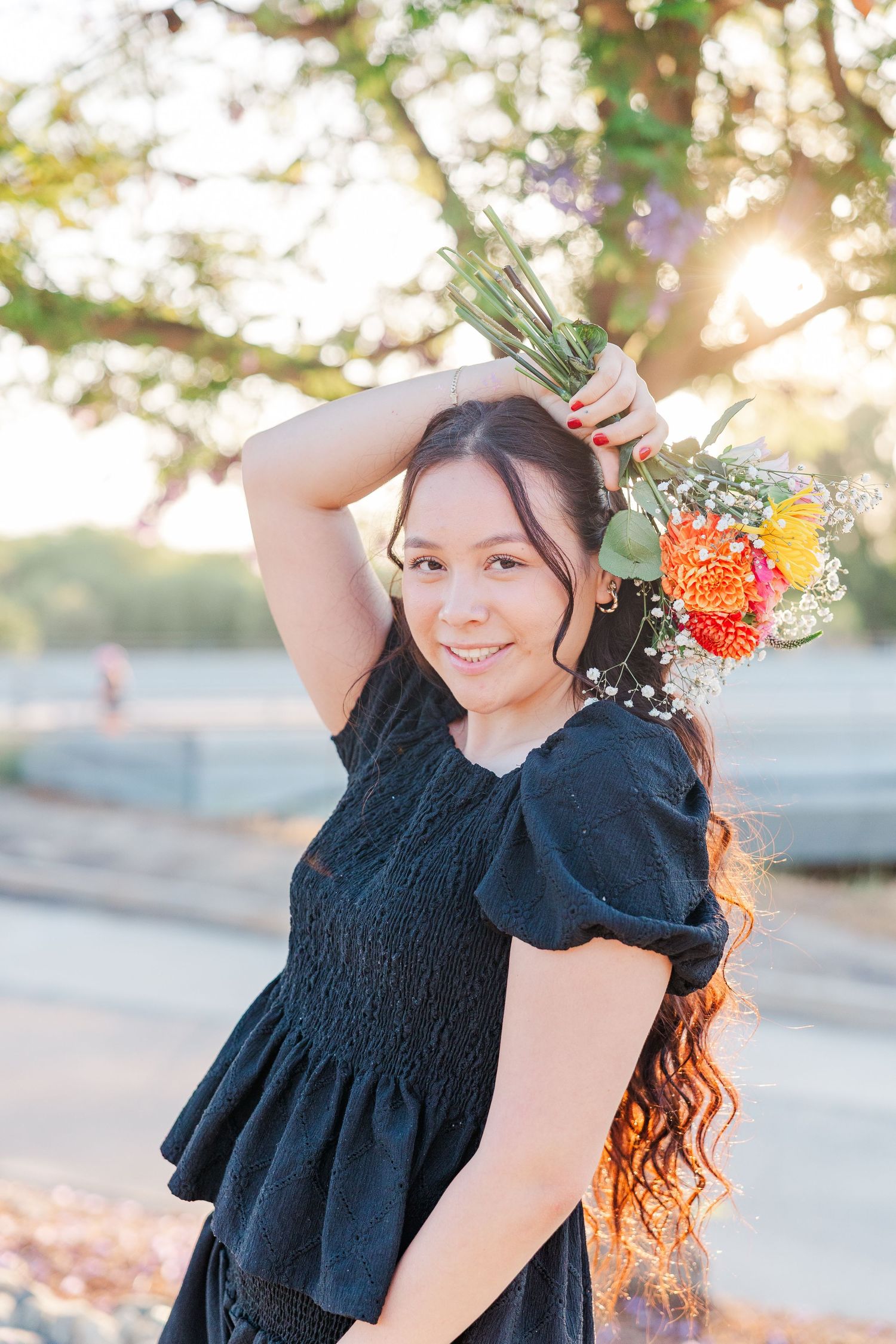 A woman in a black dress poses outdoors during golden hour with flowers in her hair.