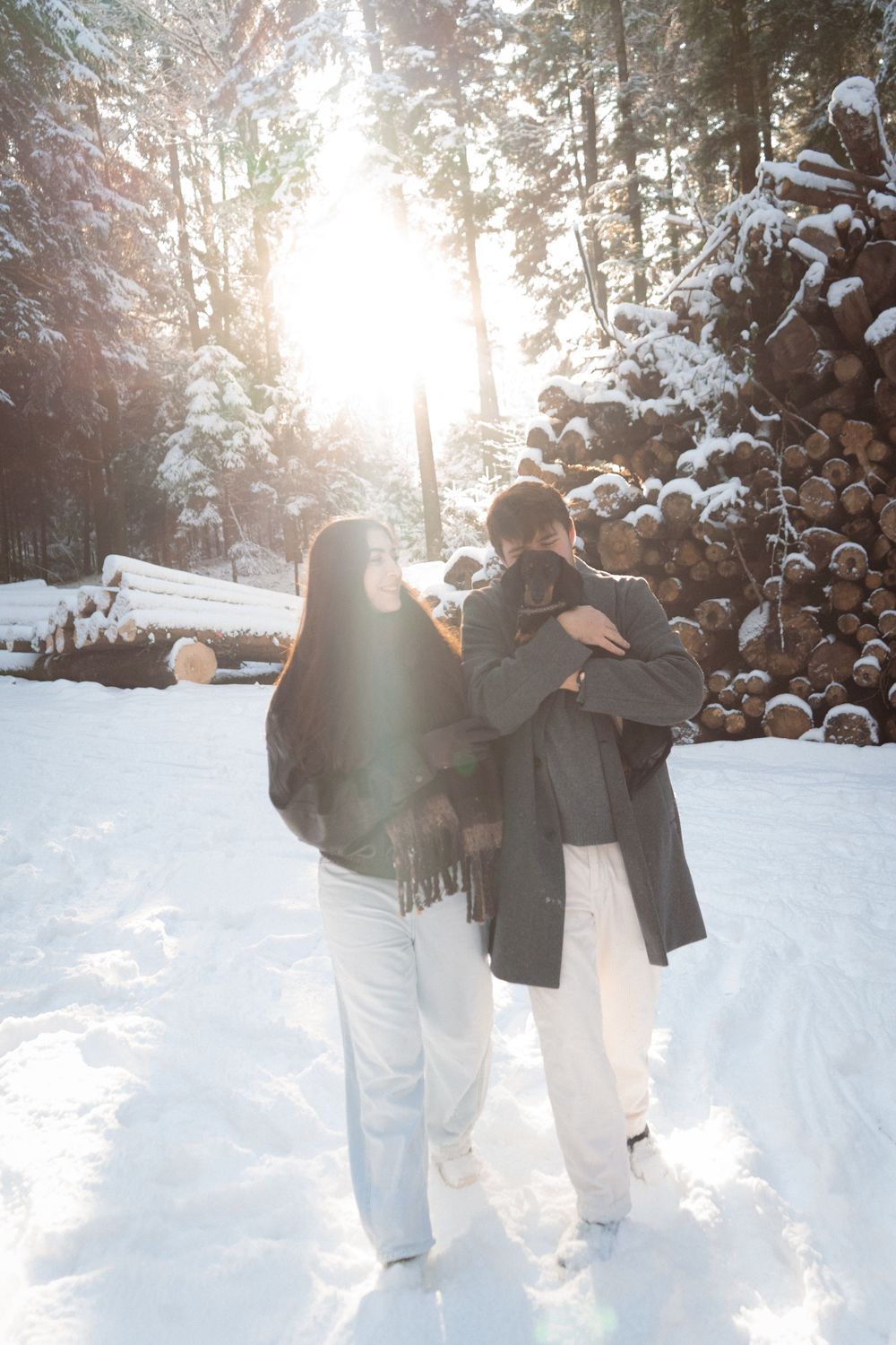 Couple walking through snowy forest path with sunlight streaming through trees.