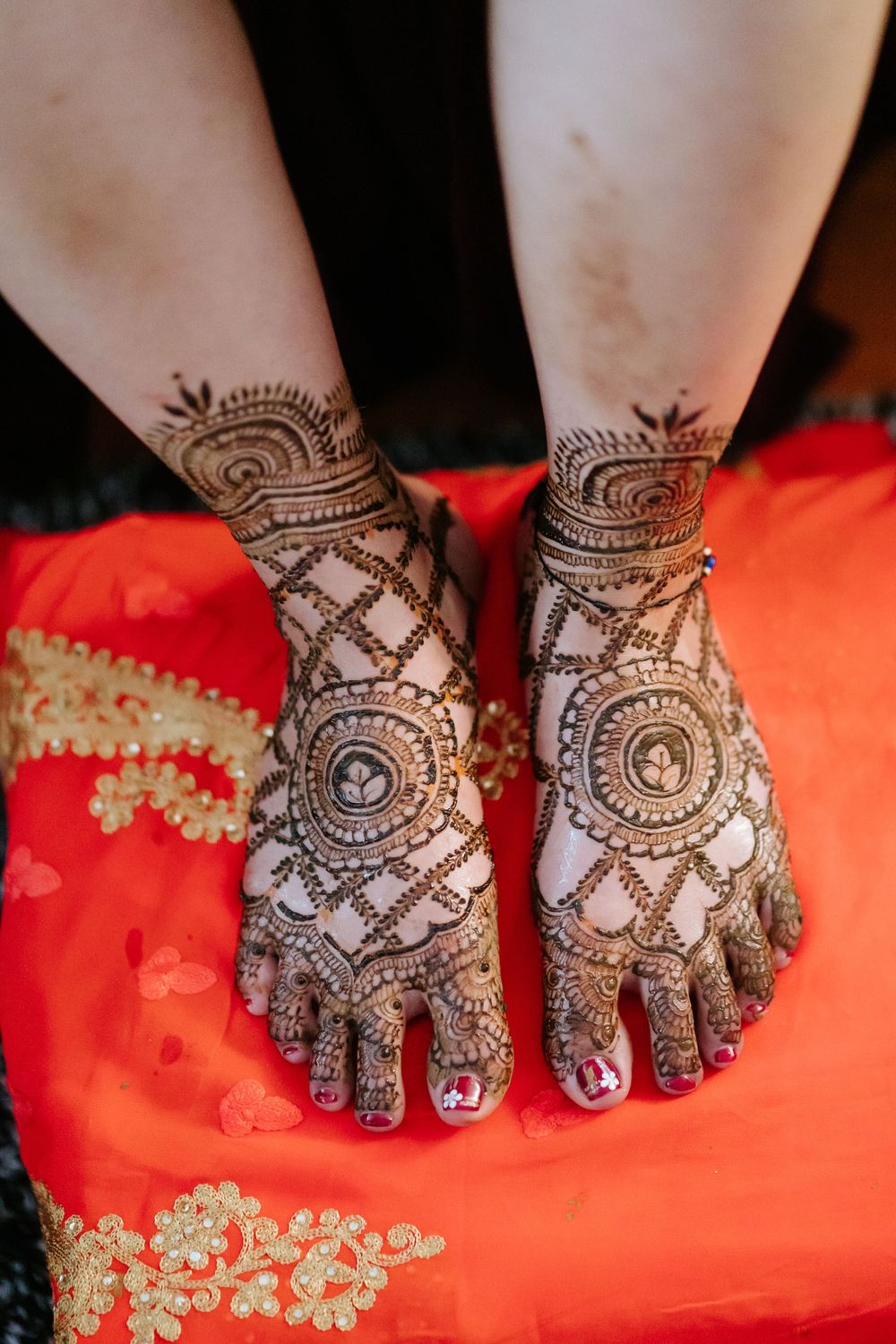 Intricate henna designs adorning hands resting on red fabric with delicate gold embroidery details.